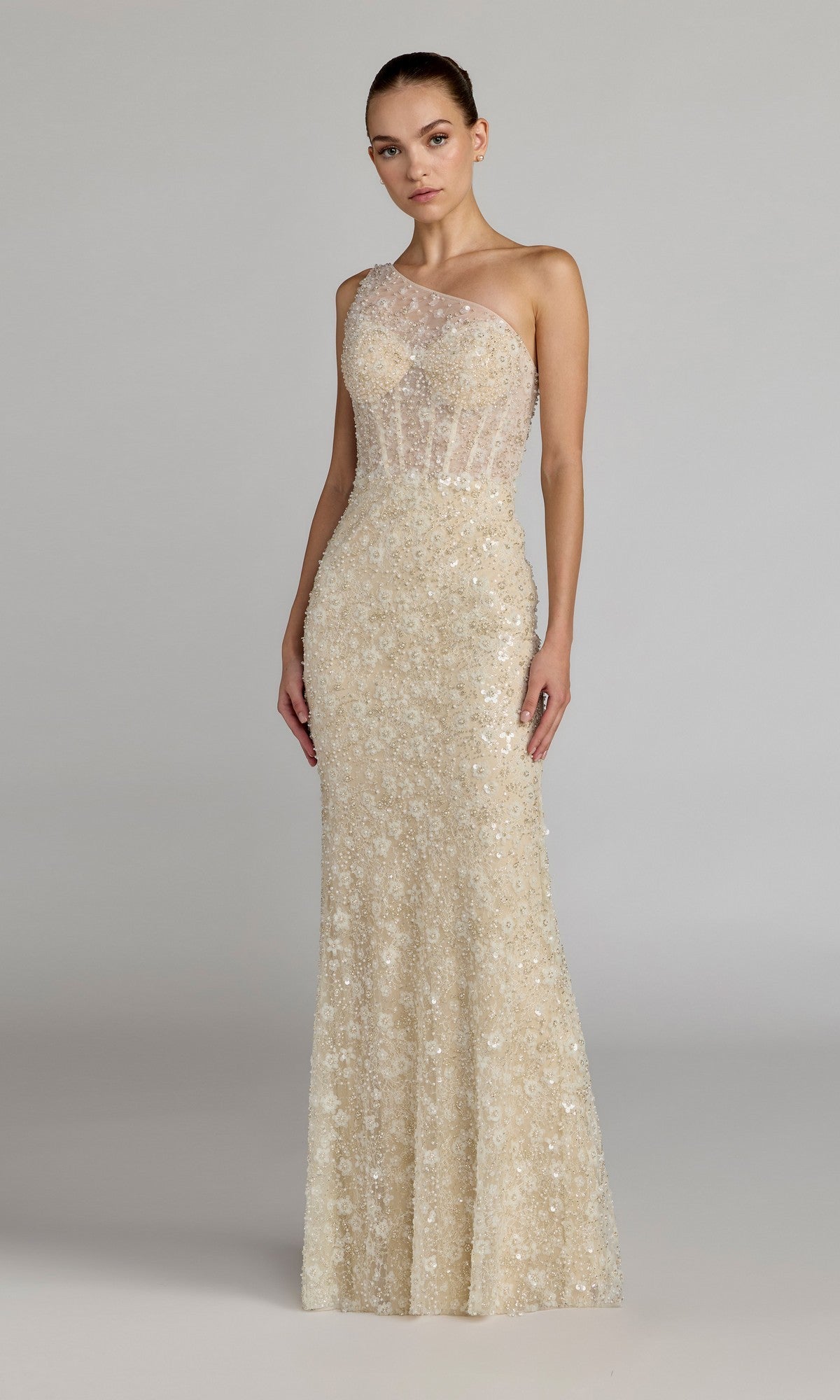 A woman stands facing forward, wearing the Long Formal Dress: Mac Duggal 21287 in Oyster Nude—a fitted, floor-length gown featuring a sheer one-shoulder design with intricate floral embellishments and a sheer bodice. The background is plain and gray.